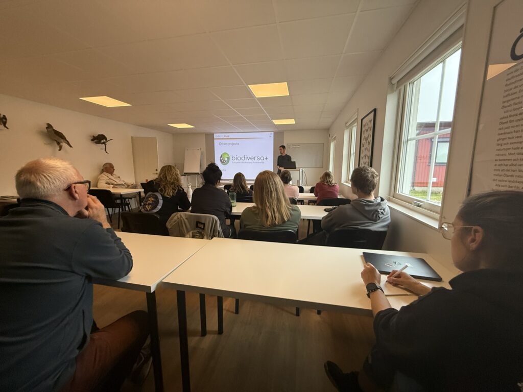 A presenter stands at the front of a classroom giving a talk on biodiversity while seated attendees listen and take notes