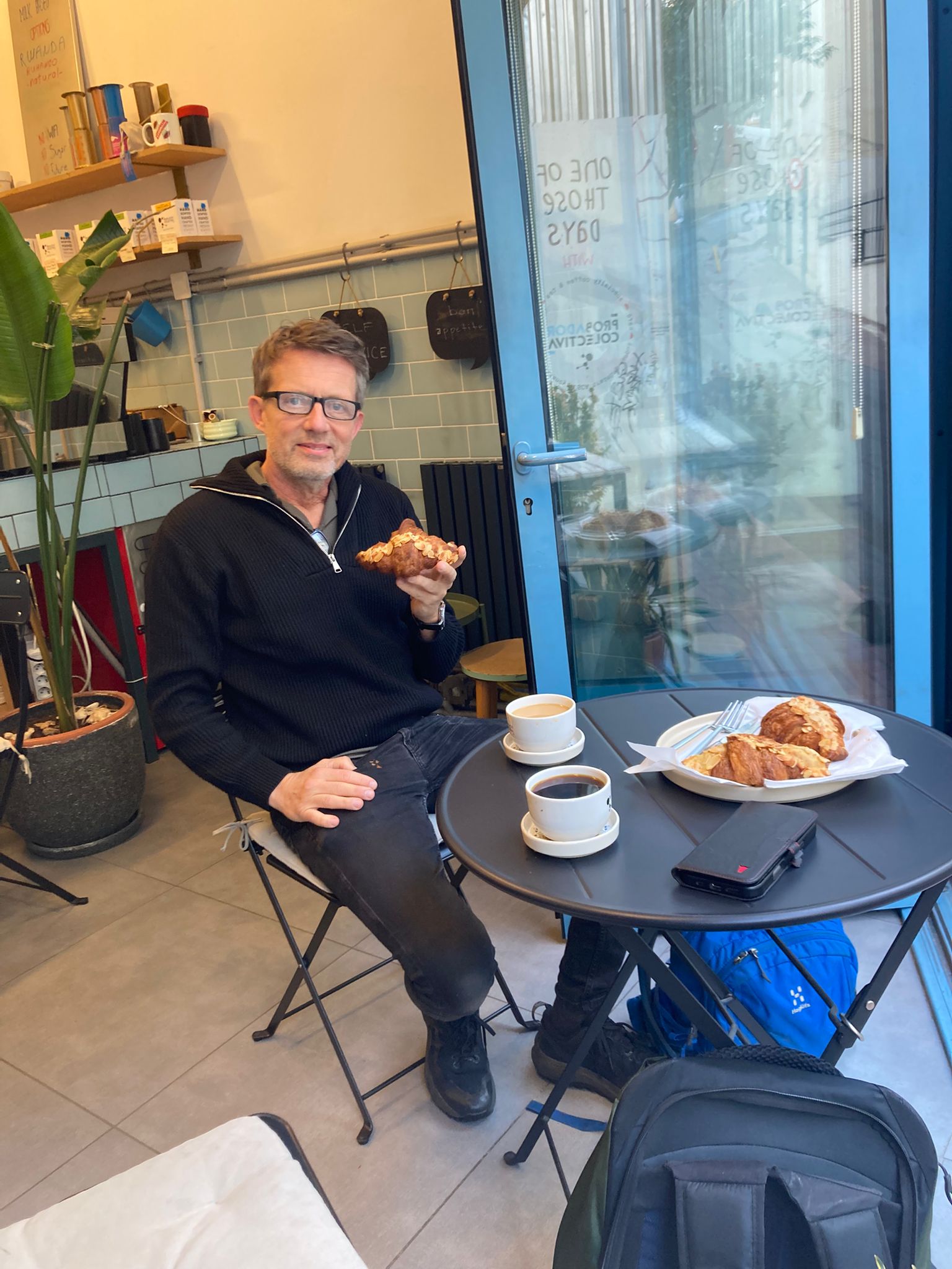 Man in glasses eating a croissant at a café table with coffee cups and pastries