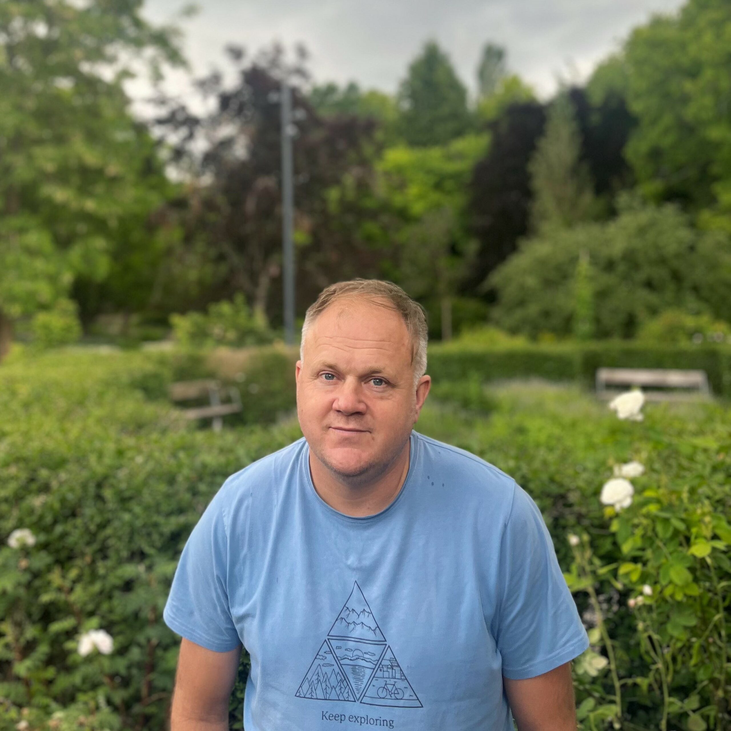 Markus Franzén, wearing a light blue t-shirt with a triangle design, standing in a garden with green hedges and coniferous t…