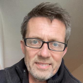 Nicklas Jansson, wearing black-framed glasses and a dark collared shirt, smiling at the camera against a neutral beige backg…
