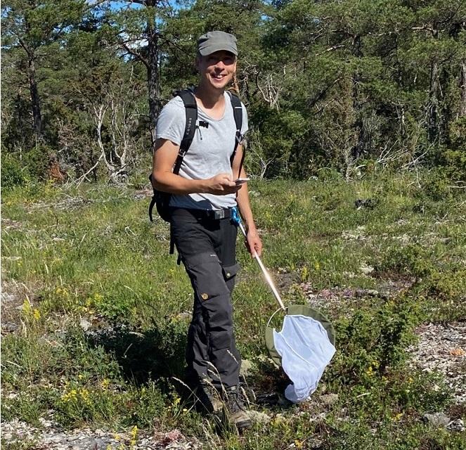 Victor Johansson, wearing a gray cap, light blue t-shirt, and black pants, holding a net while standing in a grassy woodland…