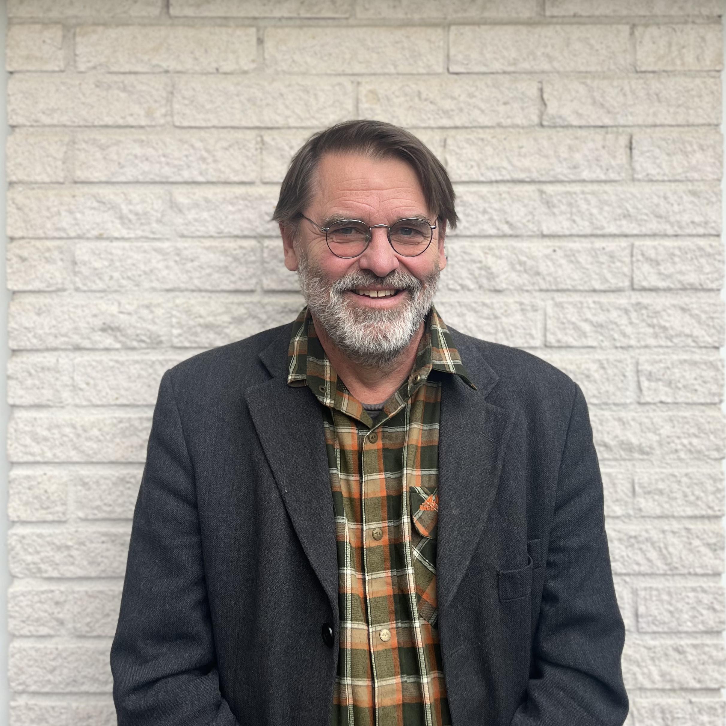 Per Milberg, wearing glasses and a dark blazer over a plaid shirt, smiling at the camera in front of a white brick wall