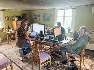 Three people work at laptops and monitors on a wooden table in a yellow-walled living room with large windows