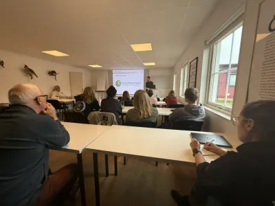 A presenter stands at the front of a classroom giving a talk on biodiversity while seated attendees listen and take notes