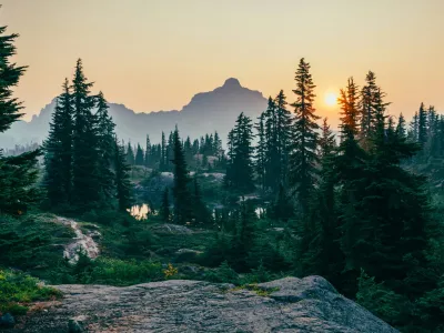 Coniferous forest with granite boulders in foreground, mountain peak and alpine lake at sunset