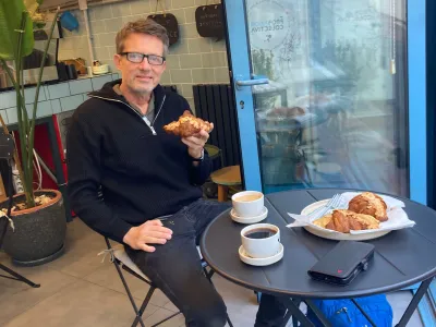 Man in glasses eating a croissant at a café table with coffee cups and pastries